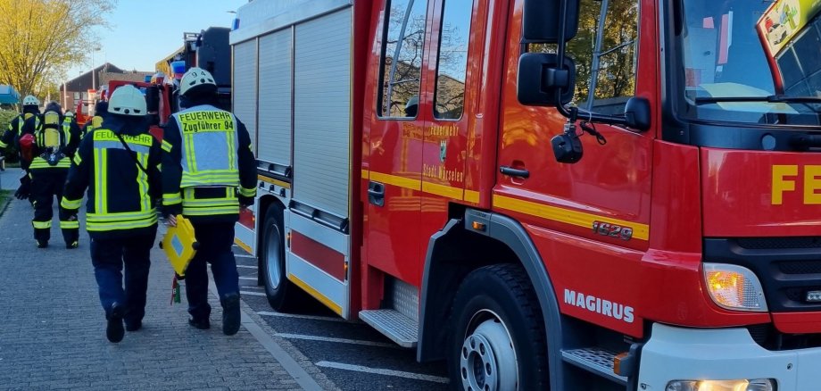 Ein Feuerwehrauto am Straßenrand und Feuerwehrleute, die von hinten zu sehen sind.