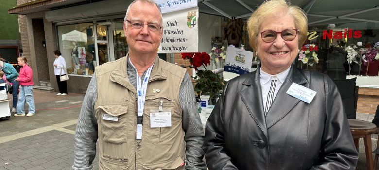 Ein älterer Herr mit grauen Haaren, beider Jacke und Brille steht auf der linken Seite. Auf der rechten Seite steht eine altere Dame mit Brille und schwarzer Lederjacke.