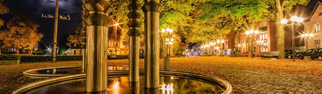 Brunnen am Lindenplatz bei Nacht. Hellerleuchtet durch die Straßenlaternen. Im Hintergrund der Maibaum der Bissener Maigesellschaft.